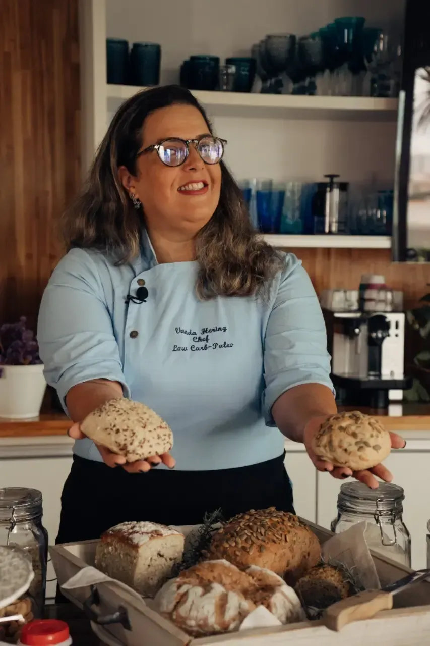 Chef Vanda Hering oferecendo dois pães low carb, com uma bandeja de pães na frente